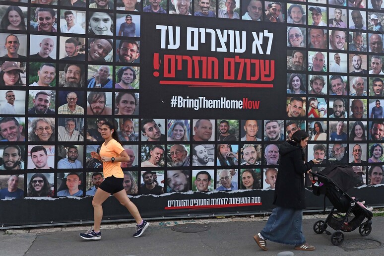 Pessoas passando por um outdoor com retratos dos reféns mantidos na Faixa de Gaza © ANSA/AFP